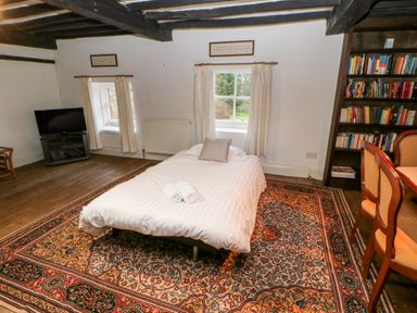 A bedroom with a bed on a patterned rug two windows a bookshelf dining chairs and a television at Eden House Edenhall near Penrith