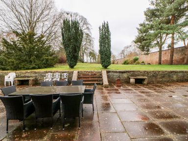 An outdoor patio area with a black table and black chairs near stone steps leading to a grassy lawn with trees and a brick wall at Eden House in Edenhall near Penrith