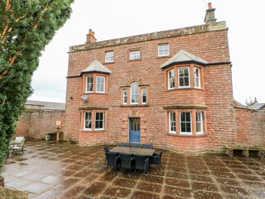 An outdoor patio with a table and chairs in front of a stone building with bay windows and a blue door at Eden House Edenhall near Penrith