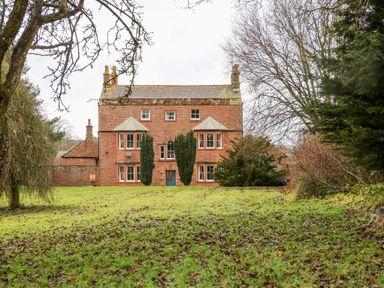A red brick house with multiple windows and a blue door surrounded by grass and trees at Eden House in Edenhall near Penrith