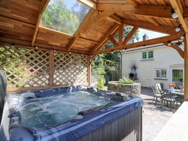 An outdoor hot tub under a wooden pergola next to a stone patio with metal table and chairs and a white house in the background at Penuwch Fach in Aberystwyth