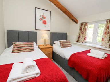 A bedroom with two single beds red throws and striped pillows a wooden bedside table with a lamp a window with floral curtains and a framed floral picture on the wall at Penuwch Fach in Aberystwyth