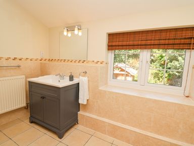 A bathroom with a gray vanity sink cabinet a mirror light fixture a window with a brown blind and beige tiled walls at Penuwch Fach in Aberystwyth