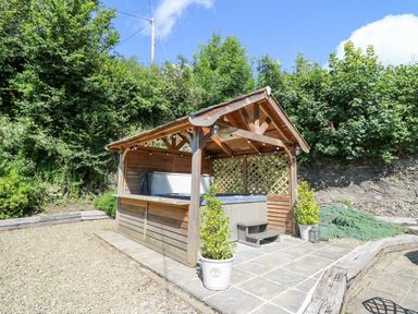 A wooden gazebo with a hot tub on a tiled patio surrounded by plants and gravel at Penuwch Fach in Aberystwyth