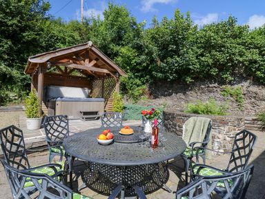 An outdoor patio with a round metal table and six chairs with cushions a hot tub under a wooden shelter and plants in the background at Penuwch Fach in Aberystwyth