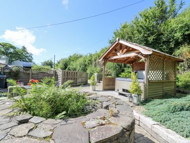 An outdoor area with a wooden gazebo covering a hot tub a stone fire pit with plants and a fenced garden at Penuwch Fach in Aberystwyth