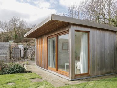 A small wooden garden building with large glass sliding doors on a stone patio surrounded by grass at Bron Heulog Cottage in Llangoed near Beaumaris