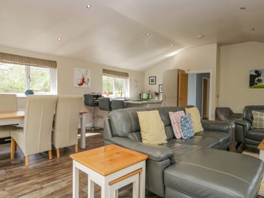 A living room with a gray sectional sofa and colorful pillows a dining table with beige chairs and bar stools at Cartmel Lodge in Allithwaite near Grange over Sands