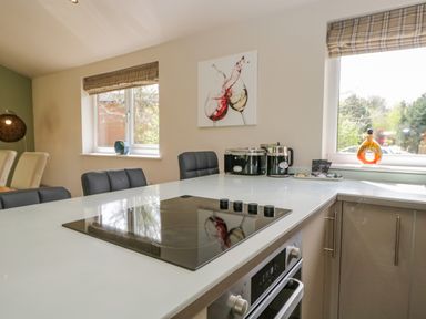 A kitchen with an electric stovetop oven and chairs by a white countertop near windows with patterned blinds at Cartmel Lodge in Allithwaite near Grange-over-Sands