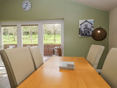 A dining area with beige chairs around a wooden table with coasters on it near glass doors showing a garden and a green wall with a clock and a picture of a building at Cartmel Lodge Allithwaite near Grange-over-Sands
