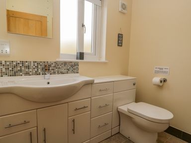 A bathroom with a white sink and cabinets a window a toilet and a toilet paper holder at Cartmel Lodge in Allithwaite near Grange-over-Sands