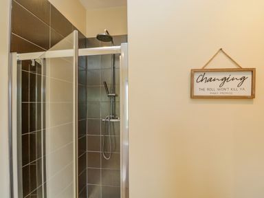 A shower with dark tiles and a glass door next to a wall with a framed sign about changing the roll at Cartmel Lodge Allithwaite near Grange-over-Sands