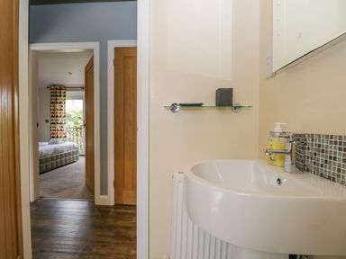 A bathroom sink with soap dispenser and a glass shelf next to an open doorway showing a bedroom with bed and patterned curtains at Cartmel Lodge in Allithwaite near Grange-over-Sands