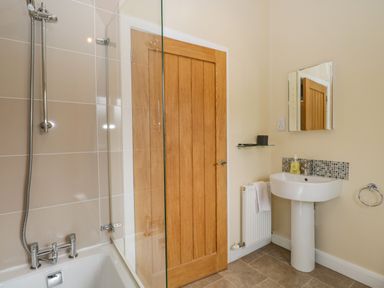 A bathroom with a bathtub and shower, wooden door, pedestal sink with mirror above, and towel hanging on radiator at Cartmel Lodge in Allithwaite near Grange-over-Sands