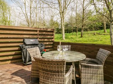 An outdoor patio with a round glass table and four wicker chairs near a wooden fence and trees at Cartmel Lodge in Allithwaite near Grange-over-Sands