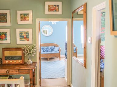 A hallway with framed pictures on the left wall, a wooden table with a vintage radio and flowers, a mirror on the right wall, leading to a sitting room with a wicker sofa and a blue rug at Craft Cottage in Beer near Seaton