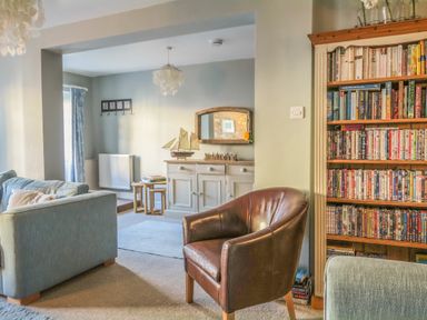 A living room with a blue sofa a brown leather chair a wooden bookshelf filled with books and DVDs and a sideboard with a model ship and mirror at Craft Cottage in Beer near Seaton