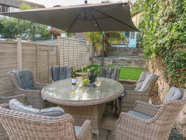 An outdoor patio area with a round glass table six wicker chairs an umbrella and greenery at Craft Cottage Beer near Seaton