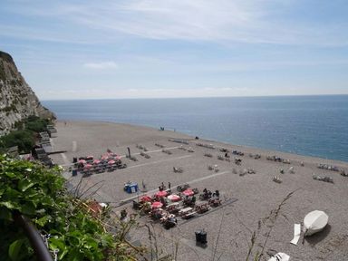 A pebble beach with sun loungers tables with red umbrellas and a covered boat at Craft Cottage Beer near Seaton