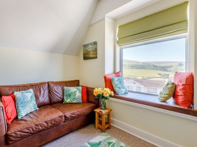 A sitting area with a brown leather sofa, cushions, a small wooden side table with flowers, and a window seat with cushions overlooking a landscape at Glenwood in Woolacombe