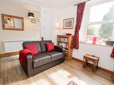A sitting area with a brown leather sofa with red cushions and blanket a wooden bookshelf with books and a lamp a window with red curtains a wooden side table and a wall mirror at Ty Gwynn in Tanygrisiau