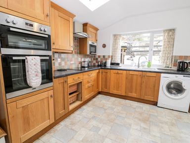 A kitchen with wooden cabinets a built in oven a microwave a washing machine and a window with curtains at Ty Gwynn in Tanygrisiau