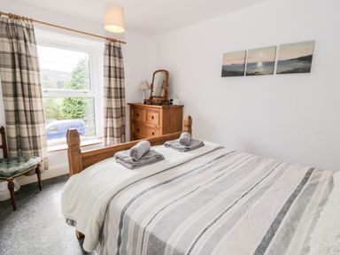A bedroom with a wooden bed, striped bedspread, two rolled towels, a wooden dresser with a mirror, a window with plaid curtains, and a chair at Ty Gwynn in Tanygrisiau