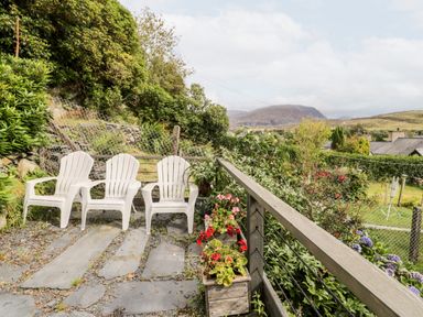 Three white plastic chairs on a stone patio with flowers in wooden planters and a garden with shrubs and mountains in the background at Ty Gwynn in Tanygrisiau