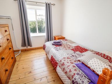 A small bedroom with a single bed covered with a patchwork quilt next to a window with gray curtains a wooden dresser and a bedside table at Marsh Cottage in North Molton