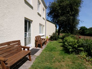 Two wooden benches along the exterior wall of a house with a garden and tree nearby at Marsh Cottage in North Molton