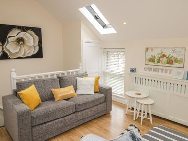 A sitting area with a grey sofa and yellow cushions near white tables and a window with blinds at The Store Rooms in Whitby