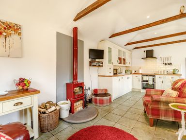 An open plan kitchen and living area with white cabinets a red wood stove and a plaid armchair at Willow Cottage in Ulrome near Skipsea