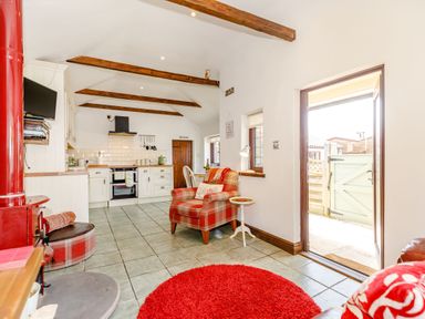 A kitchen and sitting area with wooden beams on the ceiling a red armchair a small white side table a red round rug and an open door at Willow Cottage in Ulrome near Skipsea