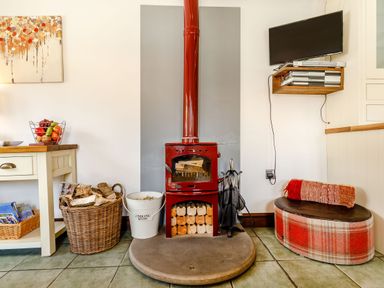 A room with a red wood-burning stove surrounded by firewood baskets a plaid cushioned seat with a rolled blanket a wall-mounted TV and a wooden table with fruit basket at Willow Cottage in Ulrome near Skipsea