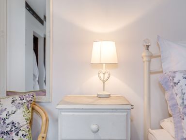 A bedside table with a lamp next to a bed and a wicker chair with floral cushions at Willow Cottage in Ulrome near Skipsea