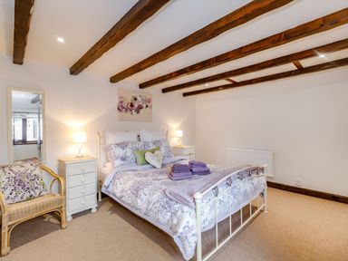 A bedroom with a double bed with floral bedding two bedside tables with lamps a wicker chair with a cushion a wall mirror and wooden ceiling beams at Willow Cottage in Ulrome near Skipsea
