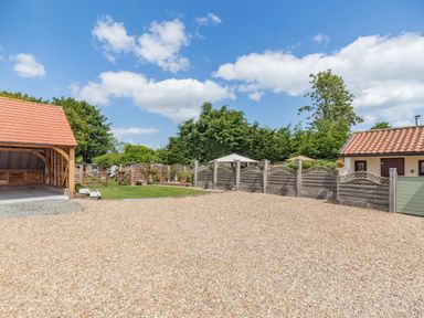 A gravel driveway with a wooden carport and fenced garden with greenery at Willow Cottage in Ulrome near Skipsea