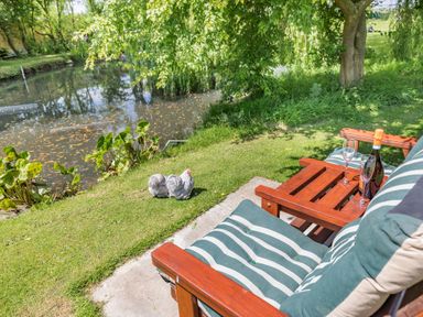 A wooden bench with striped green cushions and a bottle with two glasses near a pond and a tree at Willow Cottage in Ulrome near Skipsea