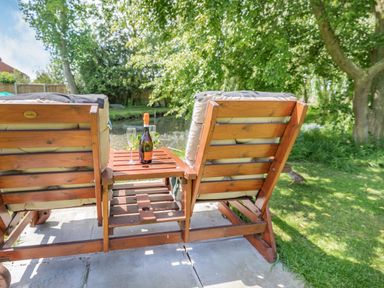 Two cushioned wooden chairs with a small table holding a bottle and two glasses by a pond in a garden at Willow Cottage in Ulrome near Skipsea