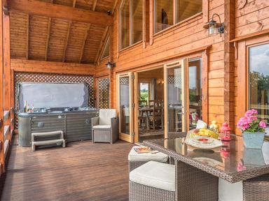 A covered wooden patio with a hot tub seating and a glass topped table with fruit and flowers at Hampton Lodge in Ellesmere