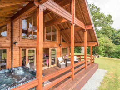 A wooden lodge with a covered porch featuring outdoor seating and a hot tub surrounded by grass and trees at Hampton Lodge in Ellesmere