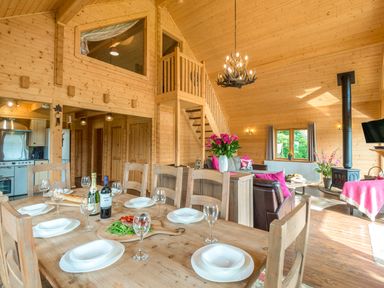 A dining table set with plates glasses and wine in a wooden cabin living room with stairs and a fireplace at Hampton Lodge in Ellesmere