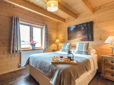 A bedroom with a bed with gray blankets and pillows a wooden tray with breakfast on the bed two bedside tables with lamps and a window with curtains and flowers at Hampton Lodge in Ellesmere