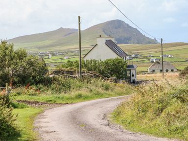 A country road curving past a house with greenery and mountains in the background at An Nead in Ballinknockane near Dingle County Kerry