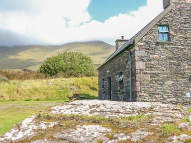 A stone house with green window frames next to a large rock and grassy hills under a partly cloudy sky at An Nead in Ballinknockane near Dingle County Kerry