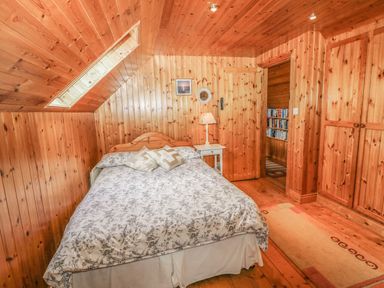 A bedroom with a bed pillows bedside table lamp wooden walls ceiling floor and a door leading to a room with bookshelves at An Nead in Ballinknockane near Dingle County Kerry