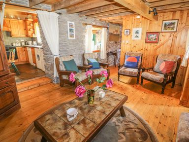 A living room with wooden floors stone and wood walls armchairs and a table with flowers and a kitchen in the background at An Nead in Ballinknockane near Dingle County Kerry