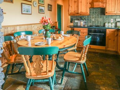 A kitchen with a round wooden table set with plates mugs and flowers six wooden chairs and wooden cabinets with tiled backsplash at An Nead Ballinknockane near Dingle County Kerry