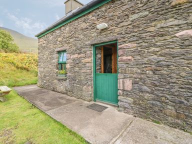 Stone cottage exterior with green door and window and concrete path and grass at An Nead in Ballinknockane near Dingle County Kerry