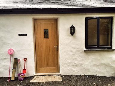 A cottage entrance with a wooden door a wall light an open black framed window and children's toys on the ground at Vine Cottage in St Ewe near Sticker